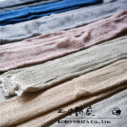 排成一列的 MOJIRI NECK’ｍ 輕柔有機棉遮陽袖套，呈現藍色、淺灰色、粉紅色混紡、淺棕色和深灰色等不同顏色。材質為輕薄的編織有機棉，觸感柔軟，部分袖套的末端帶有流蘇。