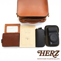 HERZ 職人手作皮革產品，包含一個棕色迷你小方包、一個棕色皮革零錢包、一個透明卡夾、一本裝飾書本，以及一個黑色的皮革筆夾和一台復古風格的相機。