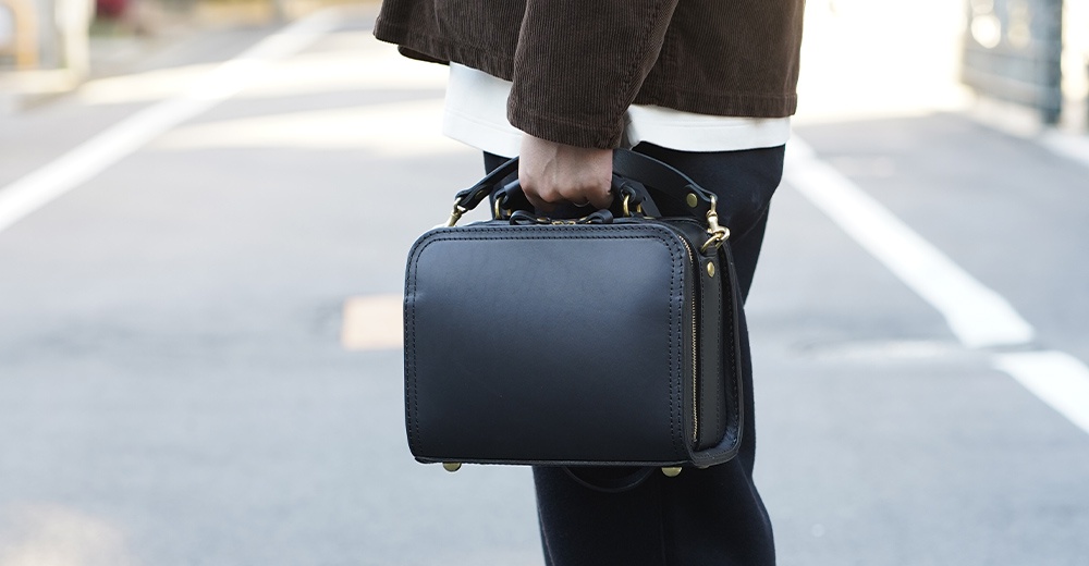 黑色皮革兩用方形單肩包，帶有頂部提把、側邊拉鍊和金色五金配件，由職人手工製作。