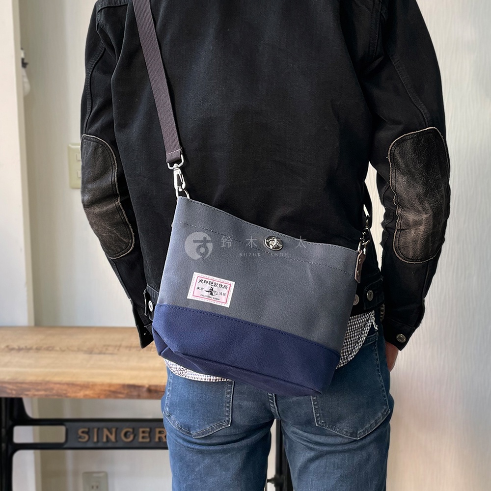 犬印純綿帆布雙色迷你側背包，採用灰色與海軍藍拼接設計，搭配深灰色肩帶，包身正面有一個印有日文文字和圖案的紅色標籤。