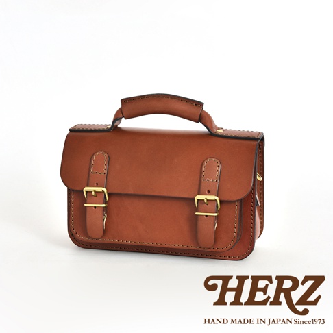 職人手作皮革雙扣兩用迷你肩背包，採用鞣製皮革製作，呈現溫潤的棕色調，包款設有雙皮帶與黃銅扣環，頂部有提把，適合肩背或手提。