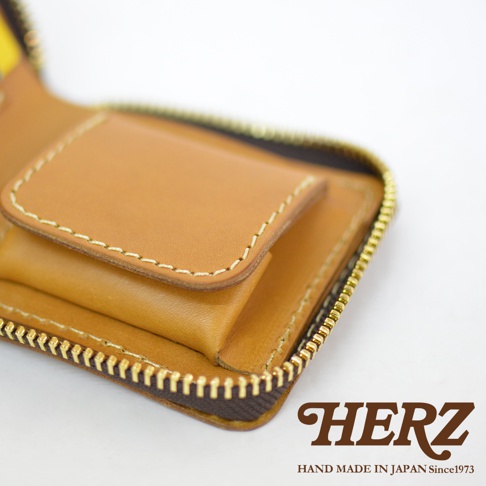 這是一張日本HERZ職人手作皮革短夾的特寫，展示了皮革溫潤的淺褐色質感與細膩的車縫工藝，包邊採用復古金色拉鍊，右下角印有HERZ品牌字樣與手作標語。