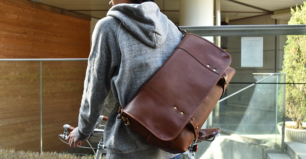 深棕色皮革復古郵差包，斜背於身穿灰色連帽衫的男子肩上，包款帶有金屬扣環和縫線細節。