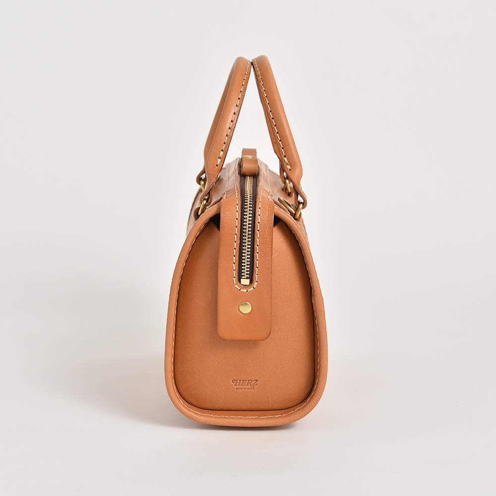 職人手作皮革迷你波士頓包，採用天然棕色皮革製作，呈現圓潤飽滿的包身輪廓。包款配有雙提把、黃銅色拉鍊及品牌標誌，整體風格簡約而精緻。