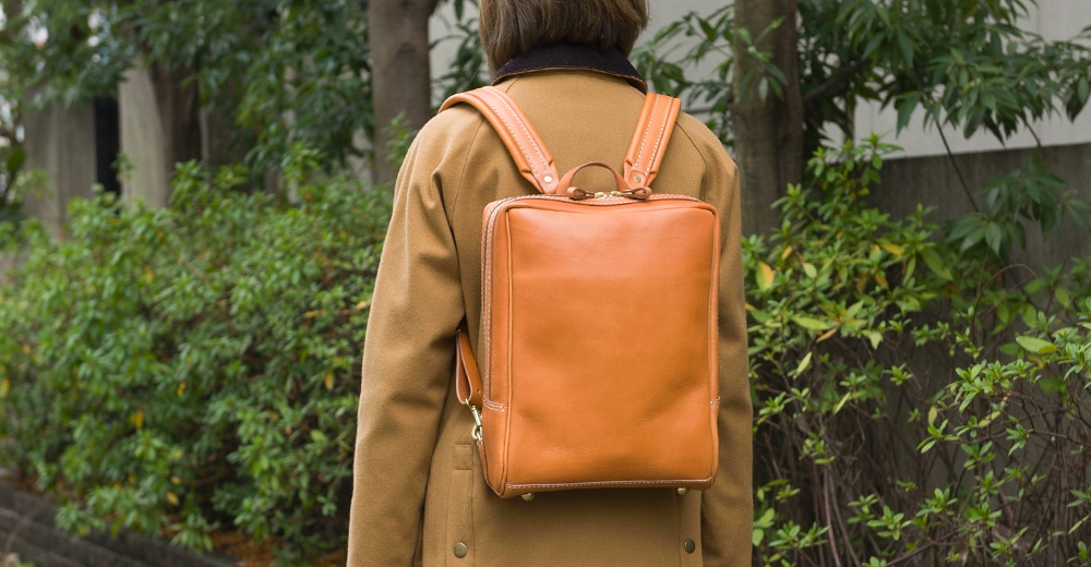 身背一個方形的棕色皮革後背包，背包有兩個肩帶、一個提把，以及金色的拉鍊。