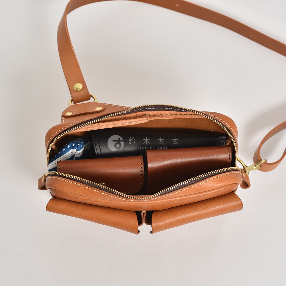 棕色皮革雙口袋斜背包，展示內部收納，包含一個黑色物品與淺棕色皮夾。