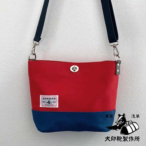 犬印純綿帆布雙色迷你側背包，主體為紅色，底部為海軍藍。包包採用帆布材質，配有可調節的深藍色肩帶，並以金屬旋轉鎖扣開合。包包正面左側有一個粉紅色標籤，印有日文文字和圖案。