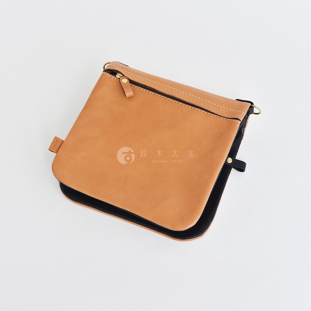 職人手作皮革翻轉雙色直式斜背包，主體為淺棕色皮革，翻蓋邊緣及內裡為黑色，具備拉鍊開合與金屬扣環。