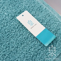 一條藍綠色長纖迷你浴巾，材質柔軟，表面有細緻的毛圈，上面放置著一張白色吊牌，印有藍色品牌標誌及「SENSHU TOWEL」字樣。
