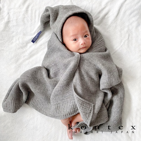 一名嬰兒被包裹在一件針織純棉的灰色連帽浴巾中，浴巾的帽子向上豎起，一條深藍色標籤從帽子一側垂下。