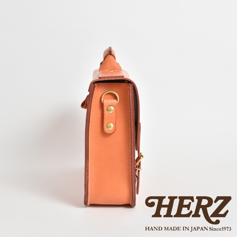 側邊視角，展示一個橙棕色皮革製成的方形公事包，包體側面有兩個銅製鉚釘和一個金屬扣環，包包上方有提把，整體為簡約設計。