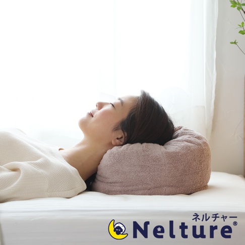 一位女子仰躺在床墊上，頭部枕於一個粉紅色、觸感類似毛巾的甜甜圈形狀枕頭，枕頭外層為絨毛材質，呈現立體環狀。