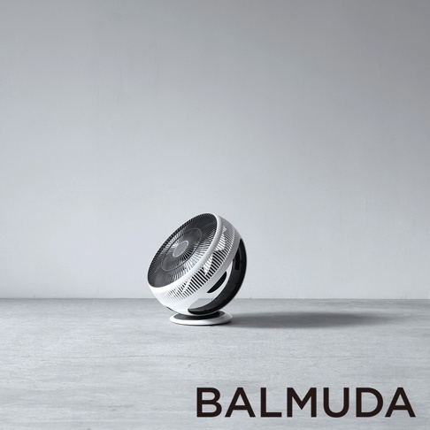 白色 BALMUDA GreenFan Cirq 循環扇，具有黑色扇葉網罩，圓形機身傾斜擺放在灰色水泥地板上，背景為白色牆壁。