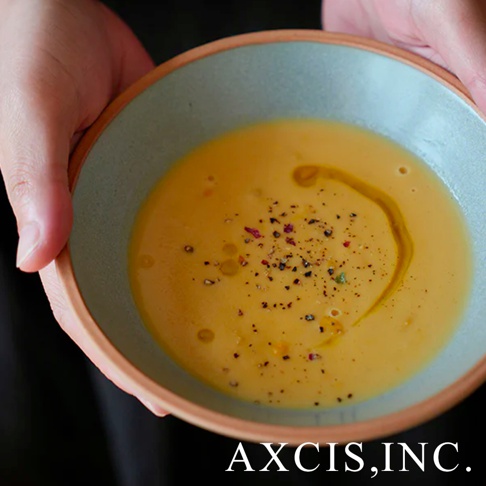 雙手捧著一個淺藍色內裡、裸色陶瓷外緣的沙拉碗，碗中盛有濃稠的南瓜濃湯，表面撒有黑胡椒、香草碎粒，並淋上少量橄欖油。