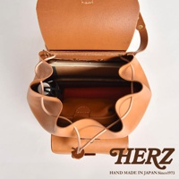 職人手作皮革經典後背包 R-4，採用天然棕色皮革製成，包身開口處有束繩設計，內部可見多層隔間，其中一個隔間放置了筆記本和紅色書籍，另一個隔間放置了淺棕色皮革化妝包。