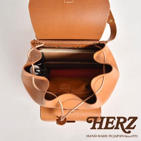 職人手作皮革經典後背包 R-4，採用天然棕色皮革製成，包身開口處有束繩設計，內部可見多層隔間，其中一個隔間放置了筆記本和紅色書籍，另一個隔間放置了淺棕色皮革化妝包。