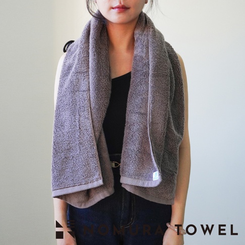 一個女人披著一條灰紫色的毛巾，她穿著黑色細肩帶上衣和藍色牛仔褲，背景是白色牆壁。