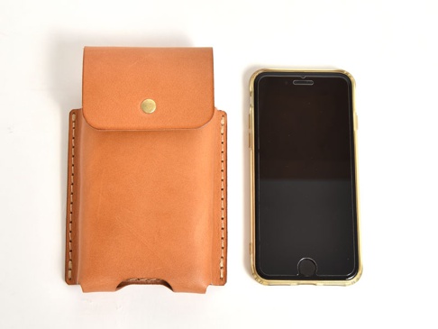 棕褐色皮革手機收納包，附有金屬按扣，旁邊擺放一部黑色螢幕、金色邊框的手機。