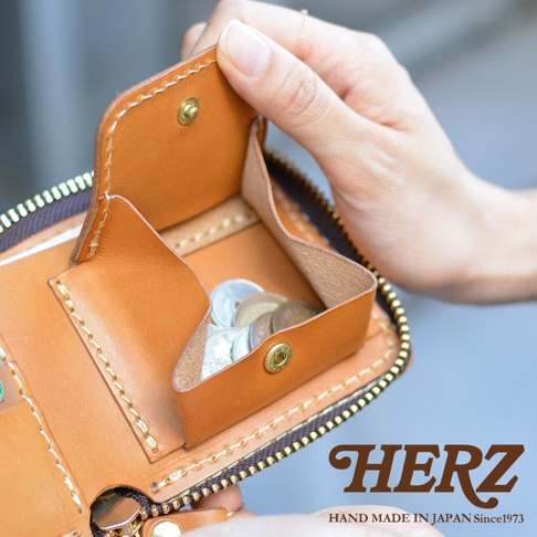 一隻手拿著淺棕色皮革製作的 HERZ 職人手作拉鍊短夾，皮夾內部結構清晰可見，設有帶有金屬扣環的皮革零錢袋，袋中裝有一些硬幣。皮夾邊緣以細膩的車縫線固定，整體呈現出高品質的日本職人手作工藝質感。