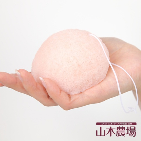 粉色蒟蒻洗臉海綿，質地細緻，帶有白色掛繩，由手部特寫展示。