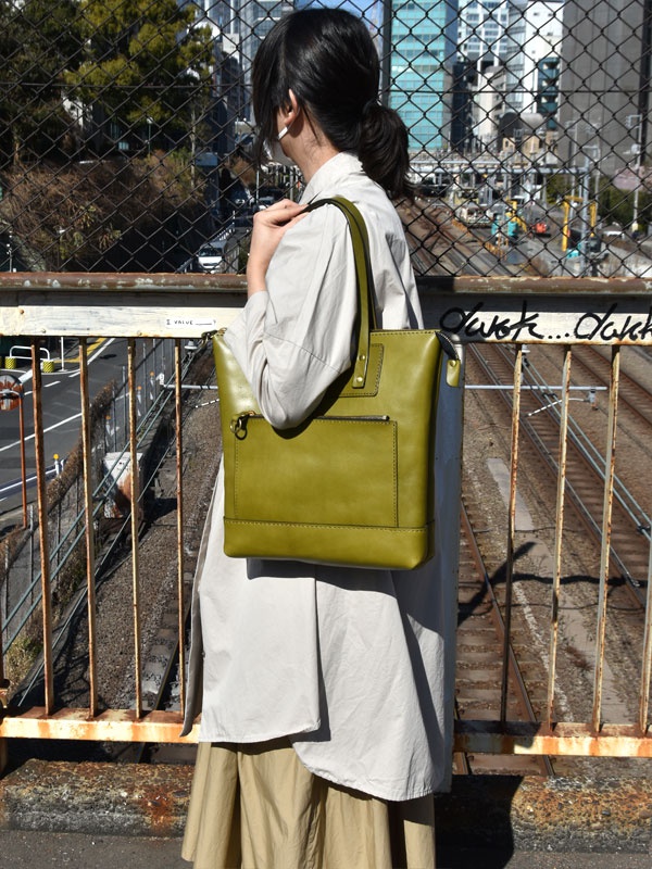 橄欖綠皮革直式拉鍊托特包，正面有一個拉鍊口袋和一個翻蓋口袋，提把上有金屬扣環，包包材質為皮革。