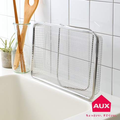 銀色金屬網格材質的伸縮瀝水籃，置於白色水槽邊緣，背景是白色磁磚牆。籃子內部有兩個金屬隔層，側邊有可伸縮的把手。旁邊有一個玻璃瓶，瓶內插著木製鍋鏟和筷子，瓶旁有一個小盆栽。