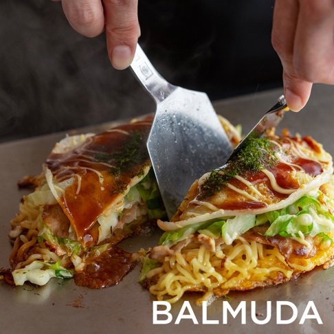 雙手使用兩支金屬鍋鏟，將加熱中的黑色電烤盤上的日式煎餅切開，煎餅內有麵條、蔬菜和醬料，表面撒有綠色粉末。