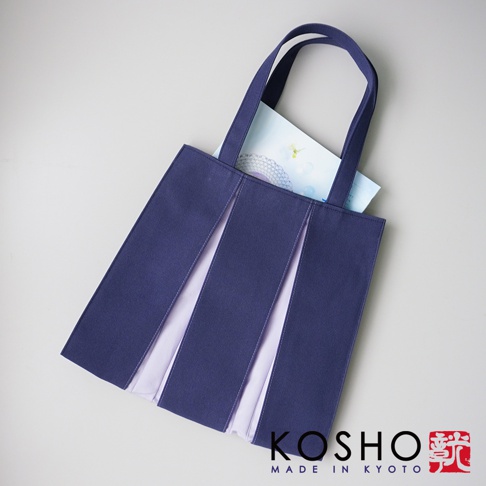 深藍色棉帆布扇形托特包，有著紫色內襯，包身有立體摺疊設計，提把為同色系棉帆布材質。