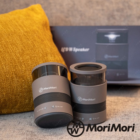 兩個圓柱形灰褐色無線藍牙音響，頂部為透明圓筒，中間有網狀格柵，品牌 Logo 標示於側。背景為黃色抱枕及印有產品名稱的深藍色包裝盒。