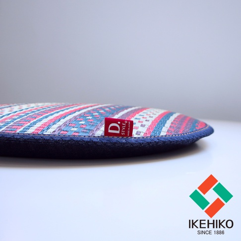 天然藺草紋織圓型坐墊，表面有粉紅色、藍色、白色相間的幾何圖紋，側邊為深藍色牛仔布包邊，並附有紅色品牌標籤。