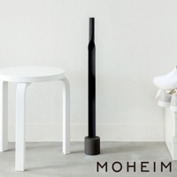 黑色金屬鞋拔，配有圓形底座，立於白色牆壁旁的灰色地板上，旁邊有一個白色凳子和一雙白色運動鞋。