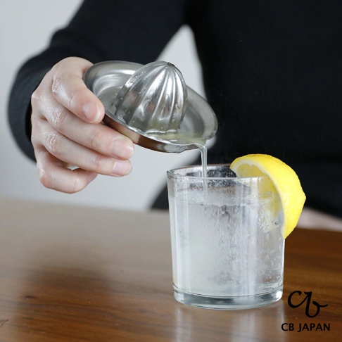 CB JAPAN 日本製不鏽鋼檸檬榨汁器正將檸檬汁擠入裝有冰塊的玻璃杯中，杯壁外側裝飾著一片檸檬。
