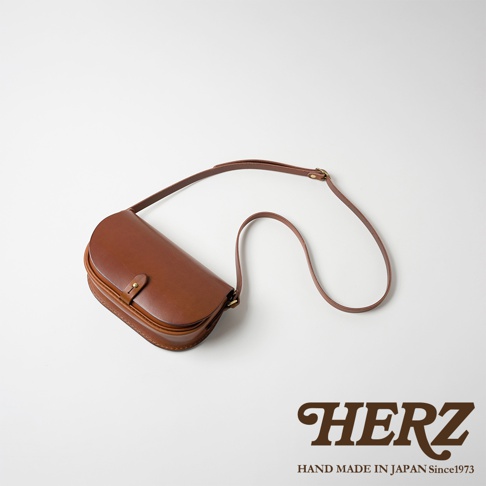 職人手作皮革迷你馬鞍包，呈現經典的半月形設計，選用光滑的棕色皮革，搭配可調節長度的細長肩帶，包身中央有一個小巧的按扣，整體散發日式職人質感。