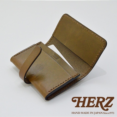 一個手工製作的 HERZ 品牌橄欖棕色皮革名片夾，呈現半開啟狀態。名片夾表面具有皮革天然質感與手工縫線細節，左側配有皮革扣帶，內側刻有 HERZ 品牌標誌與日本製造字樣，右下角標註品牌名稱與創立年份資訊。