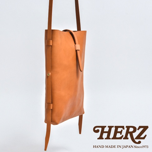 日本職人品牌HERZ手工製作的皮革直式單肩包，採用淺棕色Stare牛皮革，設計簡約纖薄。包身正面呈現側面視角，展示其獨特的皮帶釦環結構與金屬固定細節，整體外觀呈現細緻的手作質感。