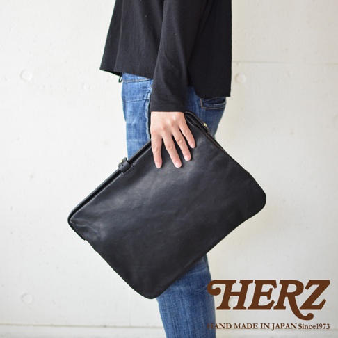 手持一個黑色職人手作皮革Organ手拿包，包體呈現柔軟的皮革質感，搭配牛仔褲與黑色上衣。