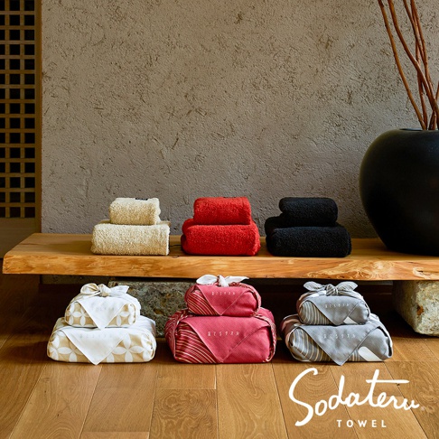 養育TOWEL wa/en 和緣紋樣風呂敷毛巾，展示於木製層架上，包含三組不同顏色（米色、紅色、黑色）堆疊的毛巾，下方地面上擺放三組以和風風呂敷包裝的毛巾禮盒，背景為粗糙紋理牆面與簡約裝飾。