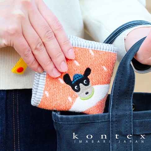 一位女性手持一條橘色系的 Kontex 今治純棉方巾，正準備將其放入深藍色的帆布托特包中。方巾上繡有一隻戴著藍色帽子、模樣可愛的動物圖案，邊緣有灰色條紋細節，整體色調柔和，展現出高品質的親膚質感與生活氣息。