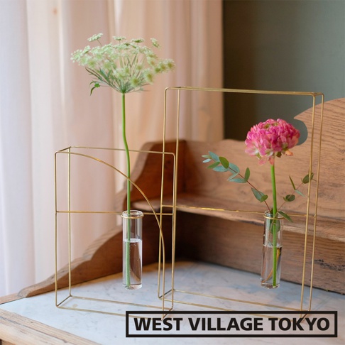 兩款簡約黃銅書本造型掛擺兩用花器，分別搭配一束綠色羽毛狀小花和一朵粉紅色花朵，置於透明玻璃試管中，展現現代設計感。