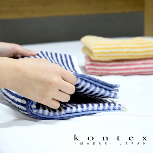 這張照片顯示一雙手正在折疊一條藍白條紋的 KONTEX 品牌棉麻迷你浴巾，背景處另外疊放著一條黃白條紋與一條粉白條紋的同系列毛巾，整體色調柔和，展現出毛巾柔軟舒適的質感。