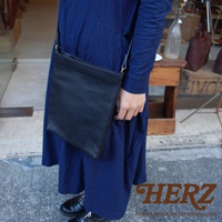 黑色皮革製的方形肩背包，垂掛於身穿深藍色連衣裙的模特兒肩上。包身簡約，材質帶有自然紋理，肩帶上可見金屬調節環。