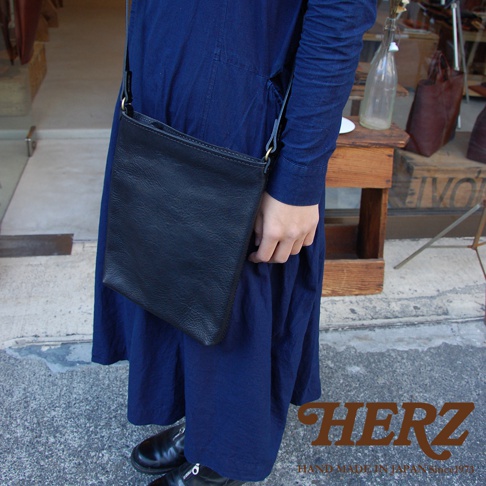 黑色皮革製的方形肩背包，垂掛於身穿深藍色連衣裙的模特兒肩上。包身簡約，材質帶有自然紋理，肩帶上可見金屬調節環。