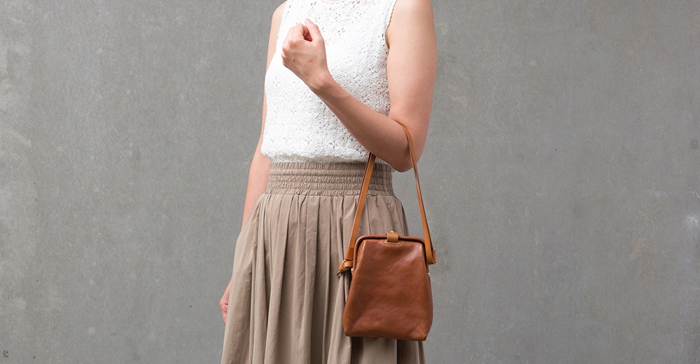 一位女性身穿白色蕾絲上衣和卡其色長裙，斜挎著一個棕色皮革迷你杜勒斯側背包，包包帶有金屬扣環。