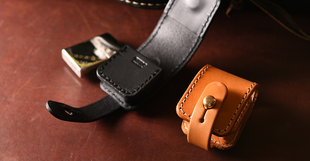 職人手作皮革原子釦型腰間小物收納包，圖片展示有黑色與原色兩款。黑色收納包帶有金屬打火機，其皮革材質與縫線清晰可見。原色收納包為方形，以皮革搭扣固定，同樣展現手工縫製的質感。