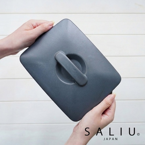 雙手持著一個深灰色的 SALIU 日本製造長方形耐熱陶製烤盤，上方蓋著同材質的平整陶蓋，頂部中央設有便於拿取的長條形手把，背景為簡約的白色橫條紋木牆。