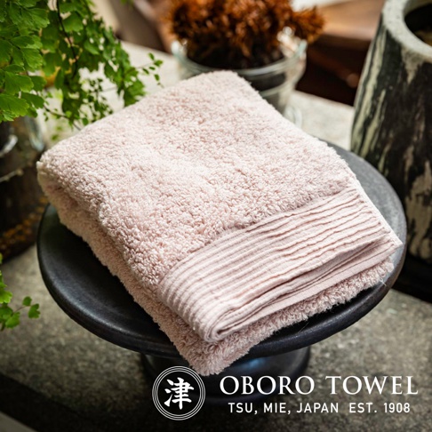 一條粉色柔軟的毛巾，表面有細緻的絨毛，邊緣有織紋裝飾，放置在黑色圓形底座上，背景有綠葉和植栽。