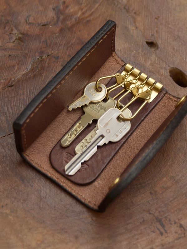 職人手作皮革鑰匙包，採用深棕色皮革，內有四個金屬掛鉤，掛有三把不同樣式的鑰匙，整體呈開放式設計，展現皮革紋理與精緻縫線。