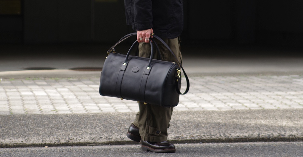 黑色皮革波士頓包，配有頂部提把、可拆卸肩帶及金色五金件，由人手提著。