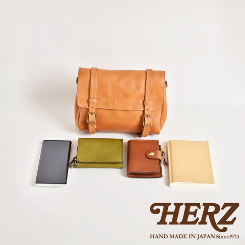 職人手作皮革水洗加工復古郵差包，配有兩個皮帶扣和皮帶。下方展示一個黑色手機、一個橄欖綠色皮革錢包、一個棕色皮革錢包和一個米色筆記本。