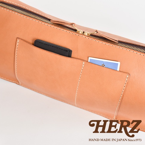 HERZ 品牌標誌，旁邊顯示 Handmade in Japan Since1973，圖片聚焦於一個淺棕色皮革包的局部特寫，拉鍊部分為金色，包身上有兩個外露的口袋，其中一個口袋裡放著一張藍色卡片。